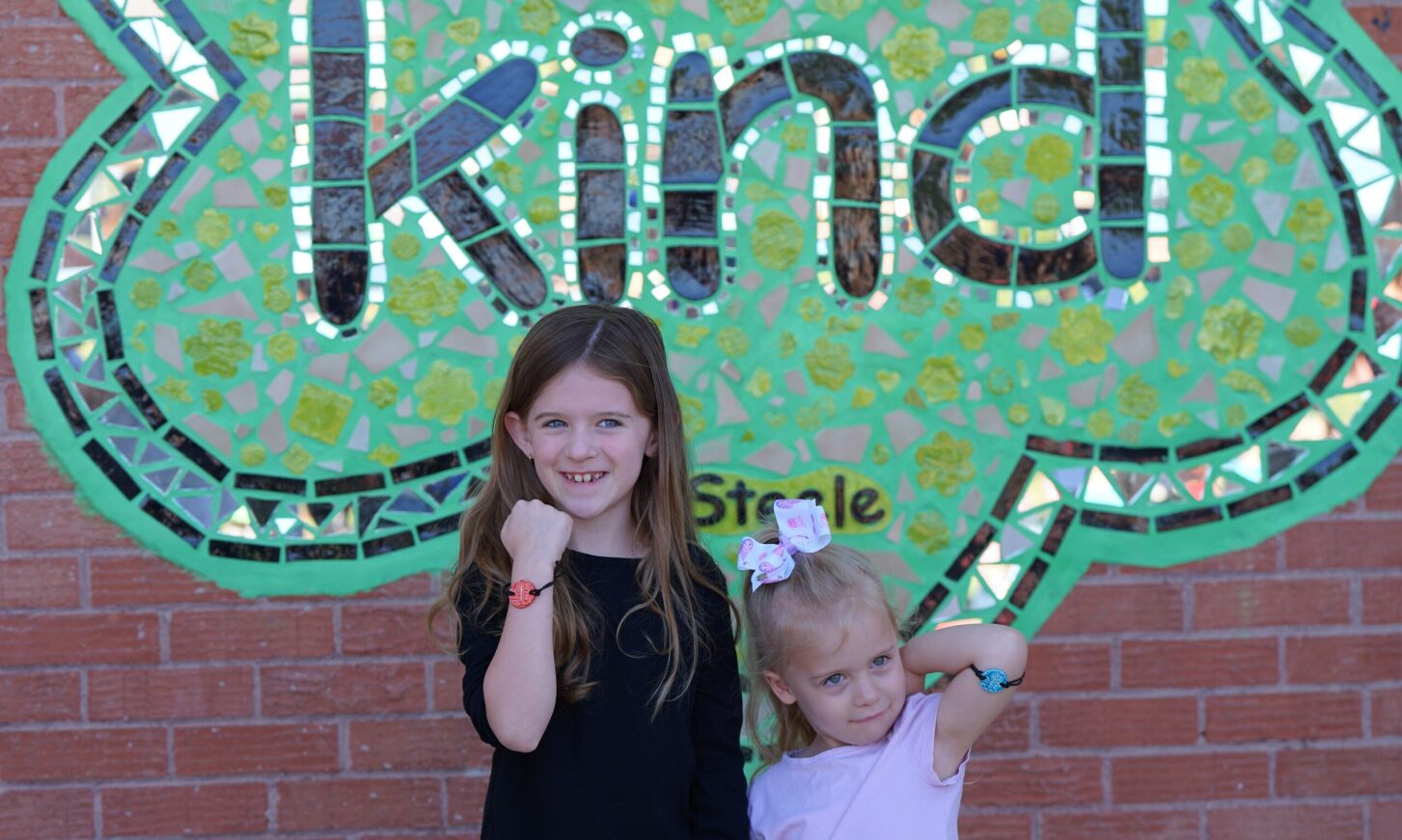 Two girls show off their bracelets in front of the Be Kind mosaic.