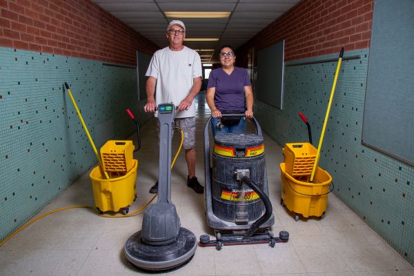 A man and woman custodian stand in the hallway with their cleaning equipment